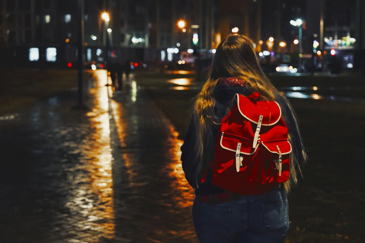 Eine junge Frau mit rotem Rucksack geht nachts allein eine nasse Straße entlang, von hinten gesehen.